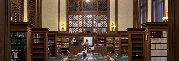 The Washington Law Library lobby, which has a dark green carpet and a brown wooden table in the middle of the room, with brown wooden bookshelves on both sides of the table. A large chandelier hangs above the front desk, where a person is talking to two clerks.