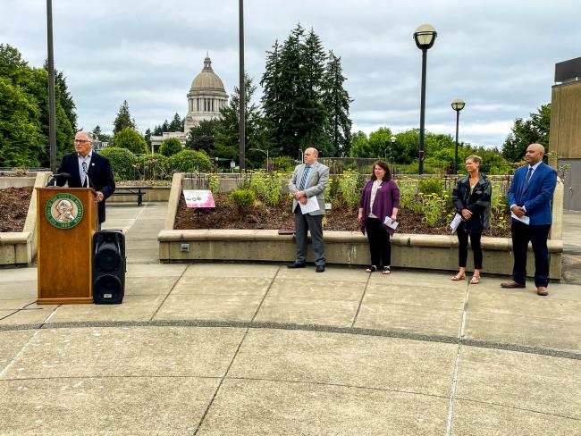 Governor Jay Inslee stands at a podium in front of the garden with the capitol building in the background