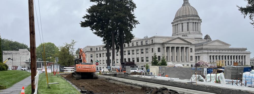 An excavator sits on a torn up section of 15th Avenue SW behind the Newhouse Building construction site with the Capitol Building in the right third of the image frame..