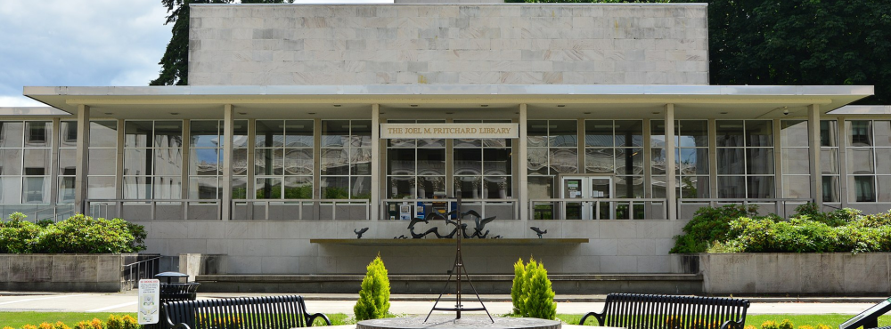 A view of the Joel Pritchard State Library's front side, which is mainly made of white stone with large glass walls. A fountain, a sundial, four metal park benches, and various green bushes are in front of the building.