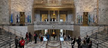 Legislative Building staff and visitors walking through the Rotunda. Some people are talking to each other, while others take pictures of the building's marble and brass features.