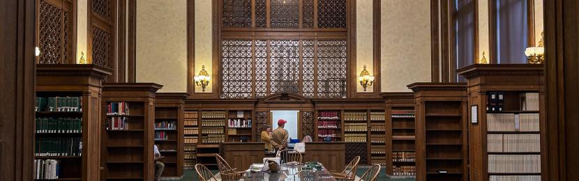 The Washington Law Library lobby, which has a dark green carpet and a brown wooden table in the middle of the room, with brown wooden bookshelves on both sides of the table. A large chandelier hangs above the front desk, where a person is talking to two clerks. 