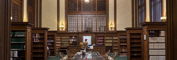 The Washington Law Library lobby, which has a dark green carpet and a brown wooden table in the middle of the room, with brown wooden bookshelves on both sides of the table. A large chandelier hangs above the front desk, where a person is talking to two clerks. 