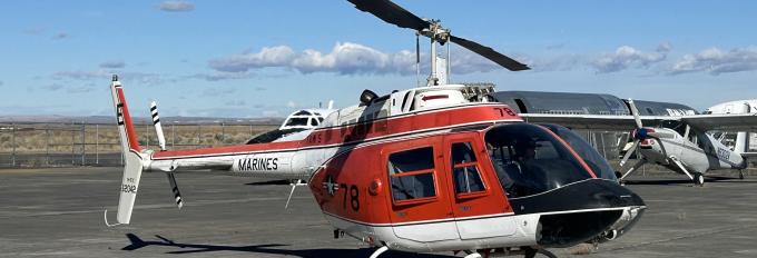 A red and white helicopter on a concrete airstrip. A small plane and two larger planes are in the background.
