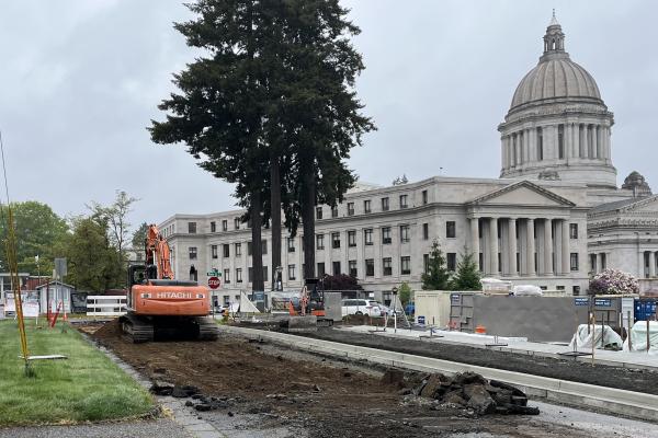 An excavator sits on a torn up section of 15th Avenue SW behind the Newhouse Building construction site with the Capitol Building in the right third of the image frame..