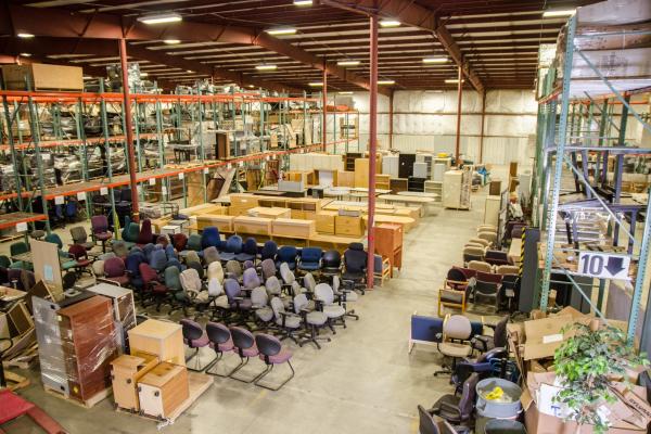 A high-angle view of various office equipment, including rolling chairs, cabinets, and desks, organized on a well-lit warehouse floor.