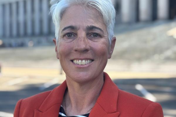 A smiling Jo Aiken stands in a red suit jacket with the Legislative Building's North steps in the background.